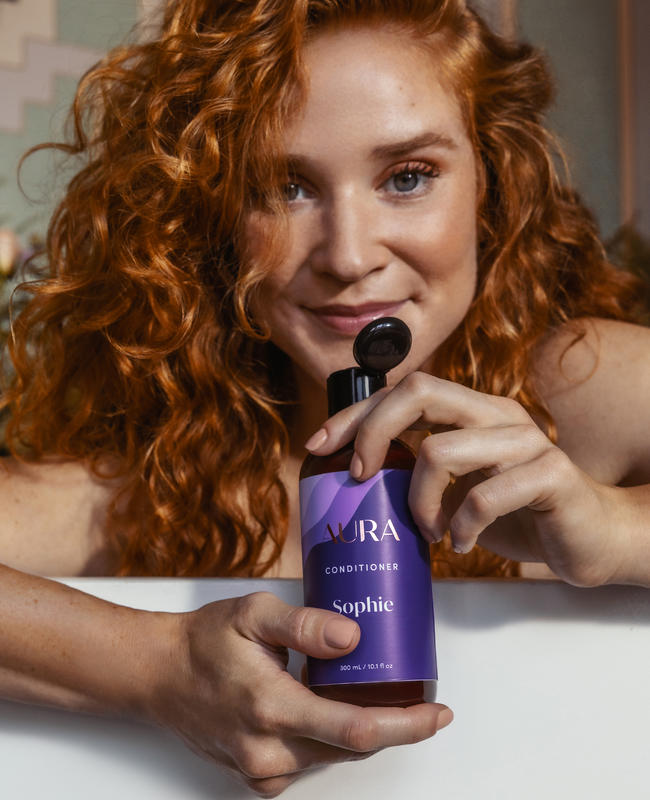 Woman with red curly hair holding her personalized AURA conditioner